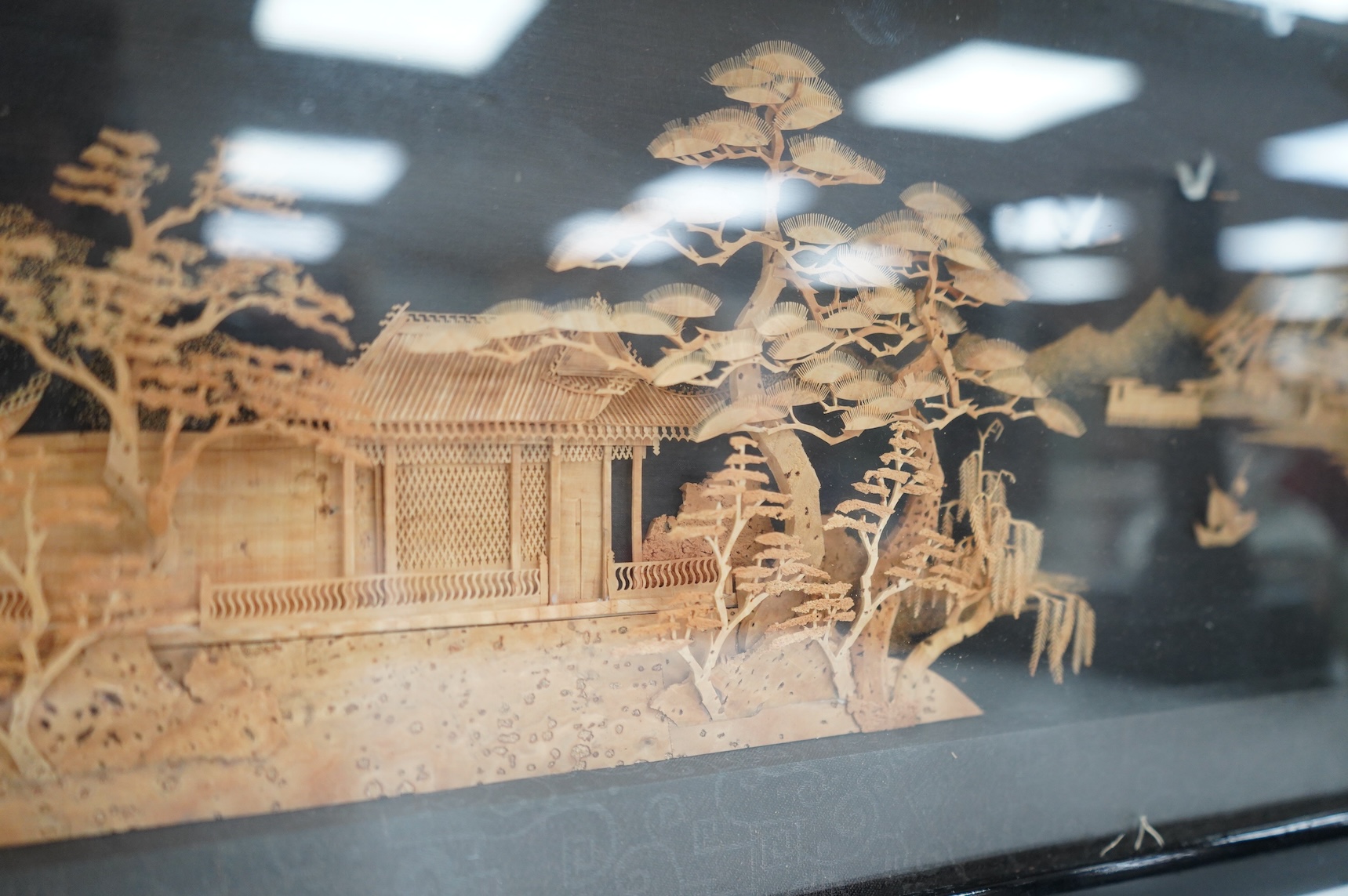 A Chinese framed, intricate relief cork landscape carving, of a Pagoda amongst trees by a river with junks and birds above., 41cm x 15cm